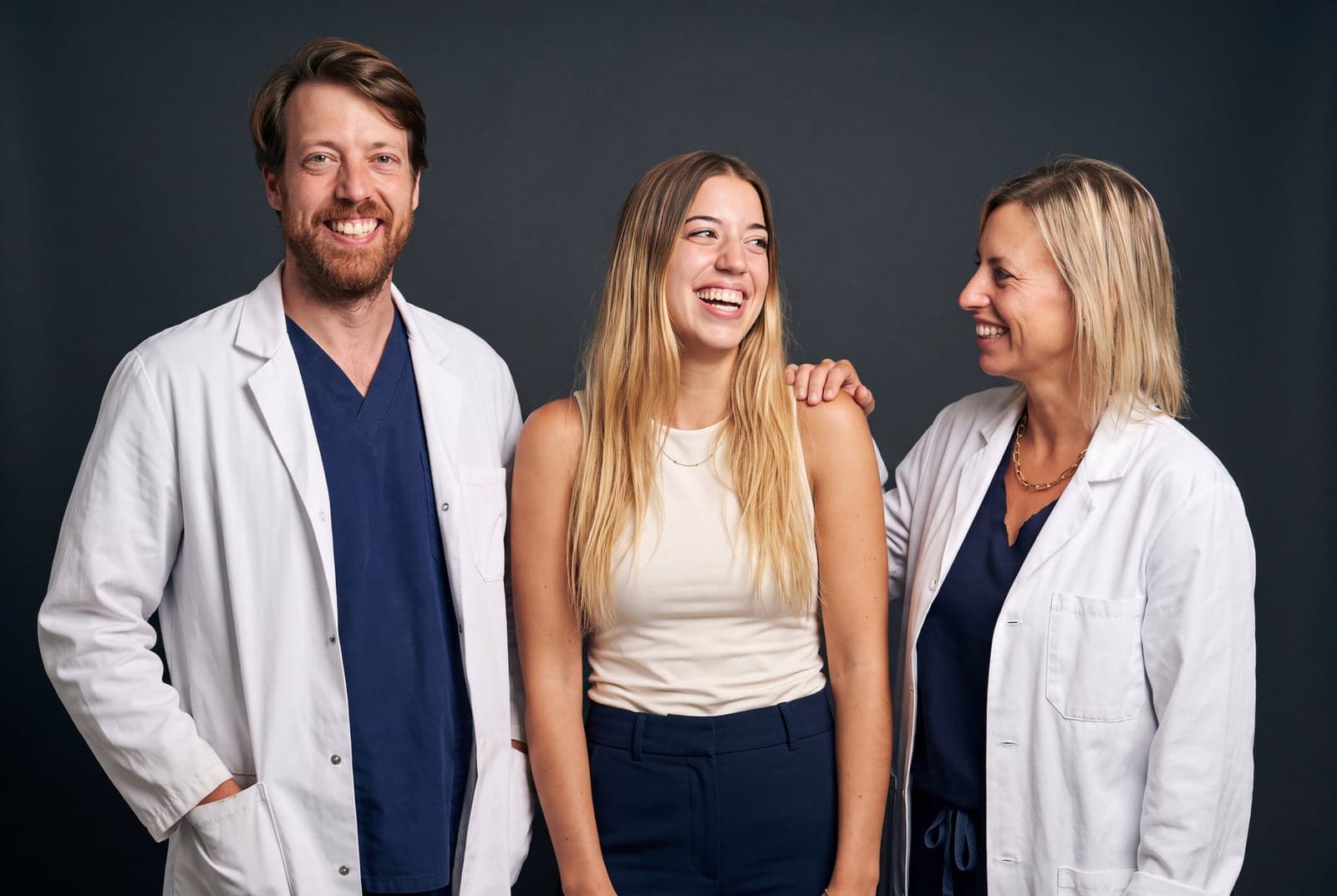 El equipo de adéu.now en AdmiraVisión Barcelona, riendo juntos sobre un fondo oscuro de estudio: el Dr. Elio Díez-Feijóo con bata blanca a la izquierda, Carla Redón en el centro y la Dra. Pilar Prats con bata blanca a la derecha